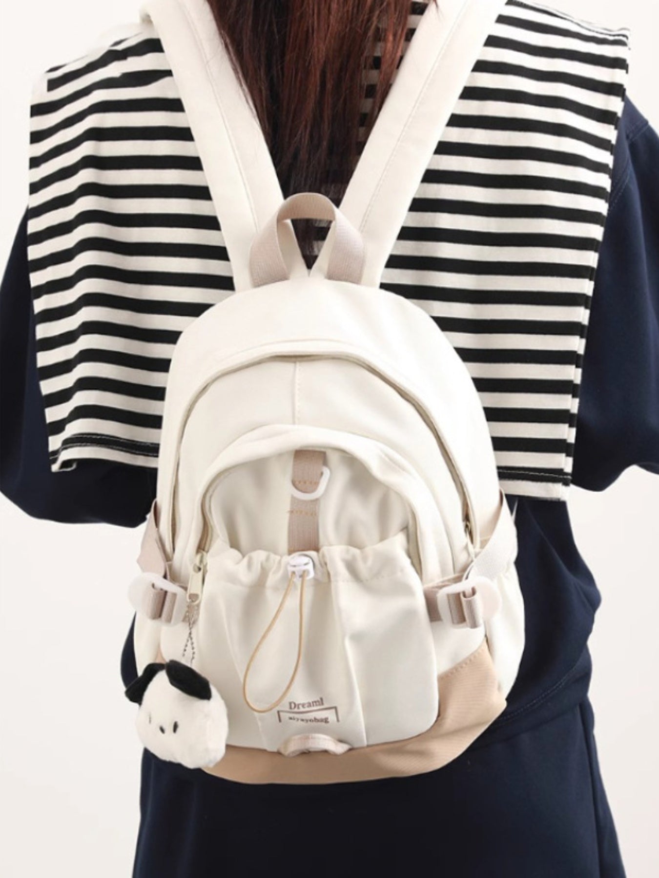White backpack with a small plush toy attached, worn by a person in a striped shirt.