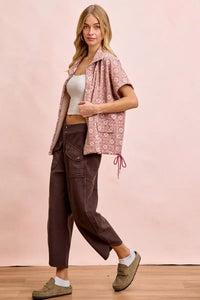 Woman wearing a pink floral shirt, white top, brown pants, and beige shoes on a pink background
