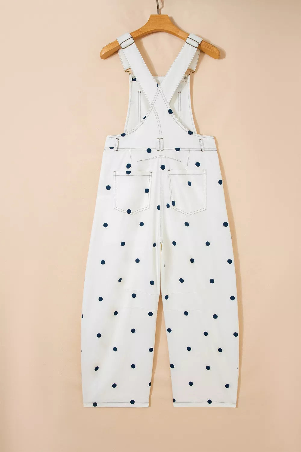 White overalls with black polka dots hanging on a wooden hanger against a beige background