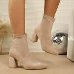 A pair of beige suede ankle boots with block heels, shown on a model's feet against a neutral indoor background with a disco ball prop.