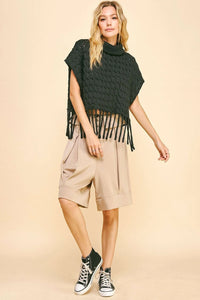 Woman wearing a black knitted poncho with fringe, beige shorts, and black high-top sneakers on a beige background
