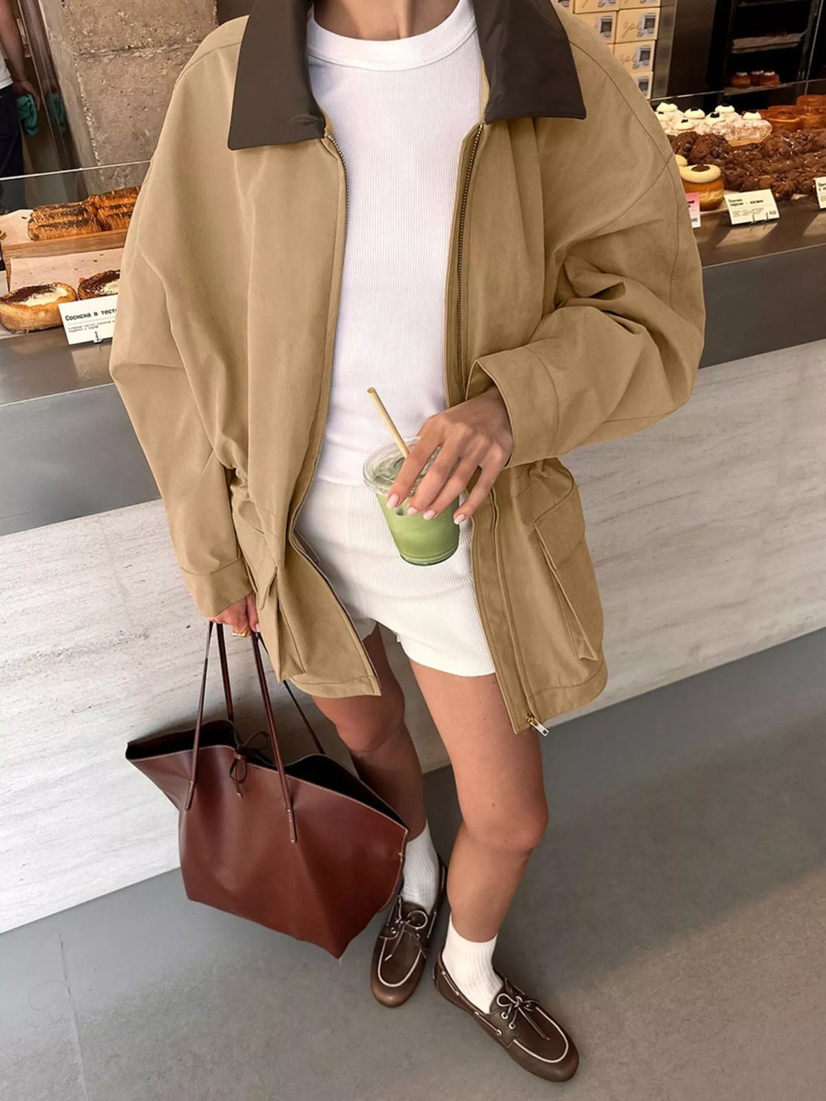 A person standing in a cafe wearing an oversized tan utility jacket with a dark brown collar, a white t-shirt, a white mini skirt, brown loafers, white socks, and carrying a brown leather tote bag.