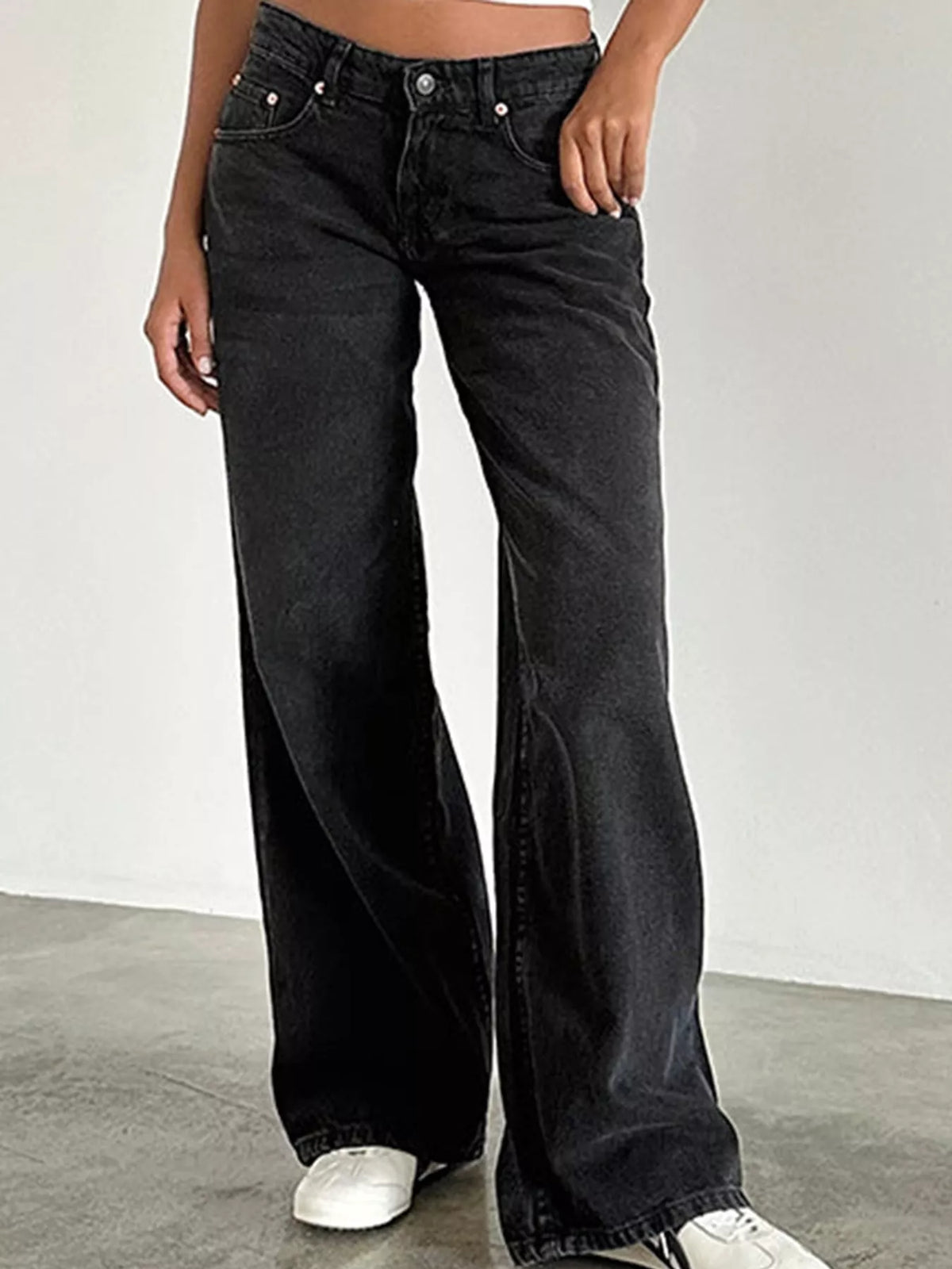 A person wearing black wide-leg denim jeans with a cropped view from the waist down, standing against a plain light gray wall and concrete floor.