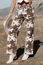A person outdoors wearing wide-leg brown-and-white cow-print pants, white ankle boots, and holding a white hat. The background shows a dry outdoor landscape with hills under natural sunlight.