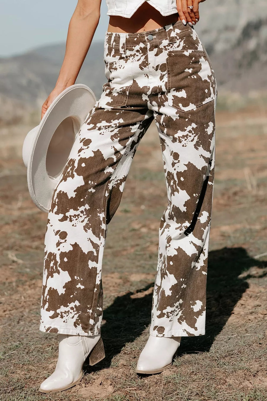 A person outdoors wearing wide-leg brown-and-white cow-print pants, white ankle boots, and holding a white hat. The background shows a dry outdoor landscape with hills under natural sunlight.