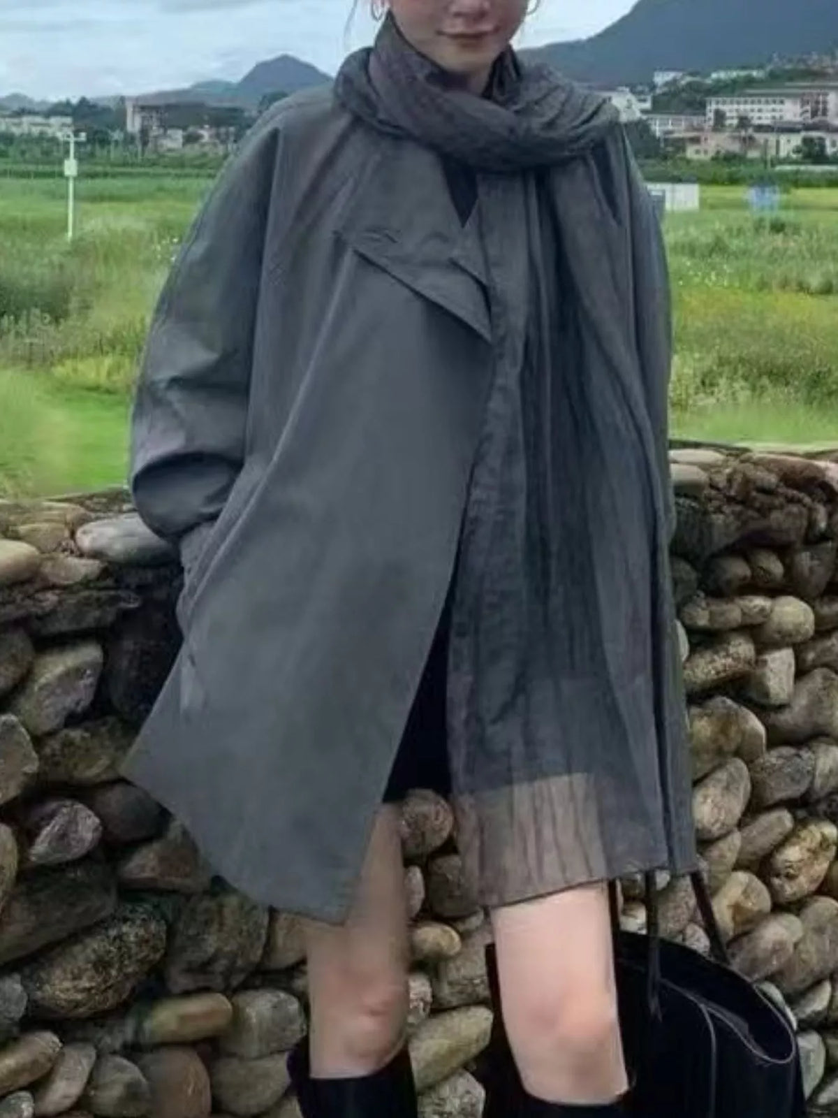 A person outdoors wearing an oversized dark gray wrap-style coat or cape with a matching scarf, standing in front of a stone wall with fields and mountains in the background.