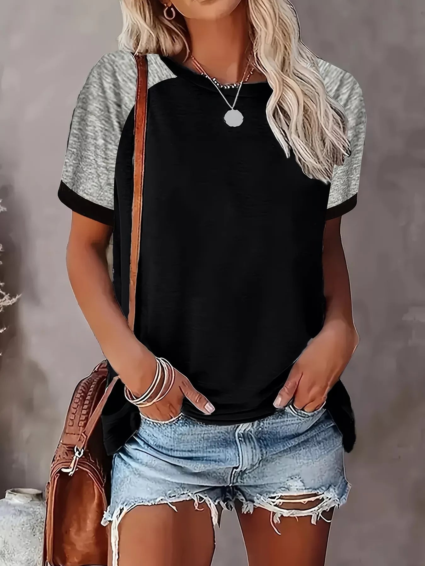 Person wearing a black and gray short-sleeve shirt with denim shorts, holding a brown bag against a gray background.