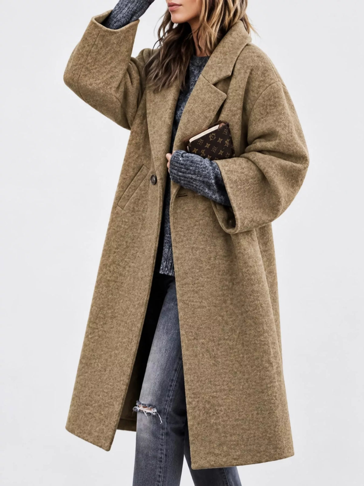 A woman wearing a long, oversized camel-colored wool coat over a gray knit sweater and distressed blue jeans, holding a brown monogram clutch against a plain light gray background.