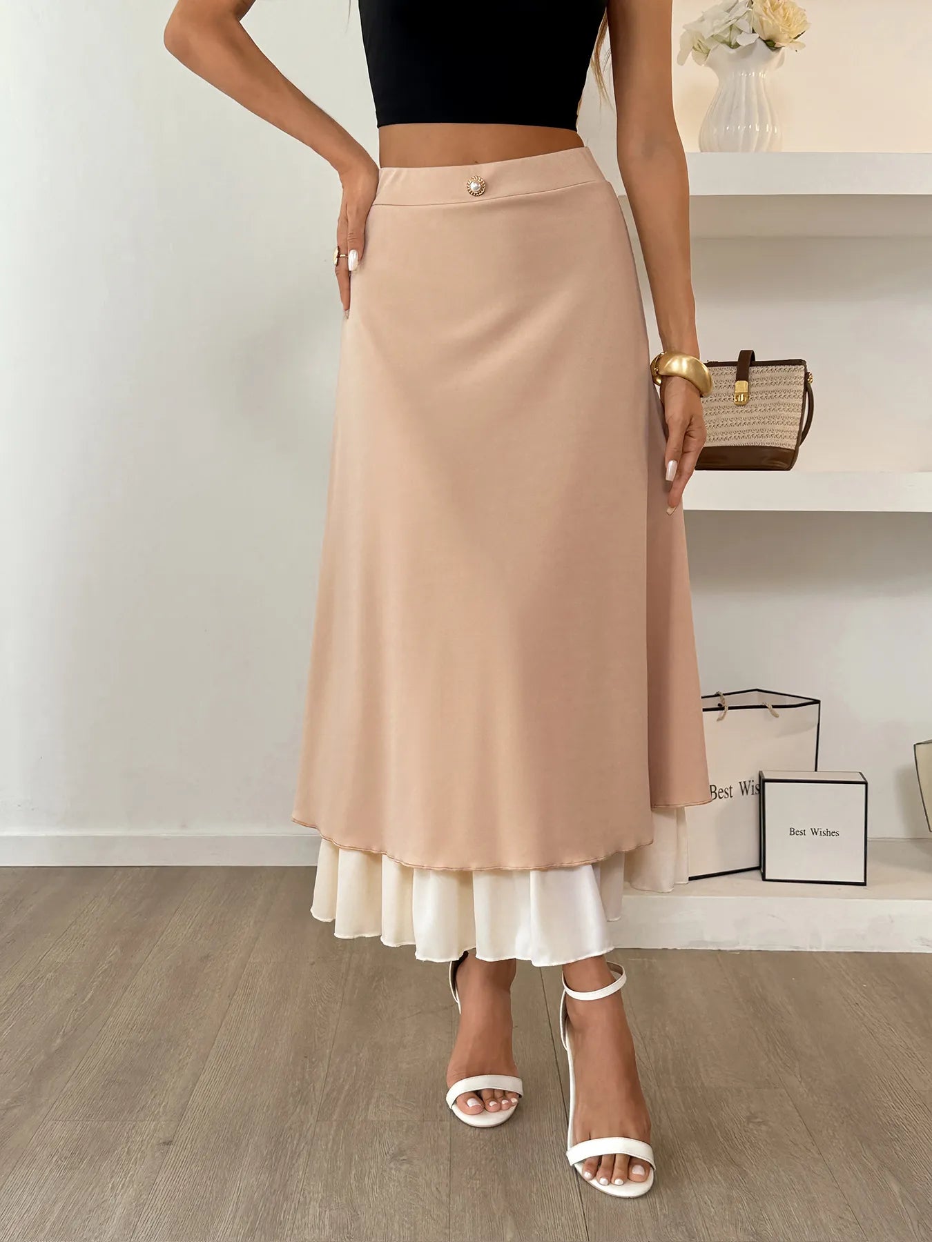 Person wearing a beige skirt with a white bottom, black top, and white sandals in a minimalistic setting.