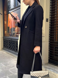 Person wearing a long black coat holding a patterned handbag on a city street.