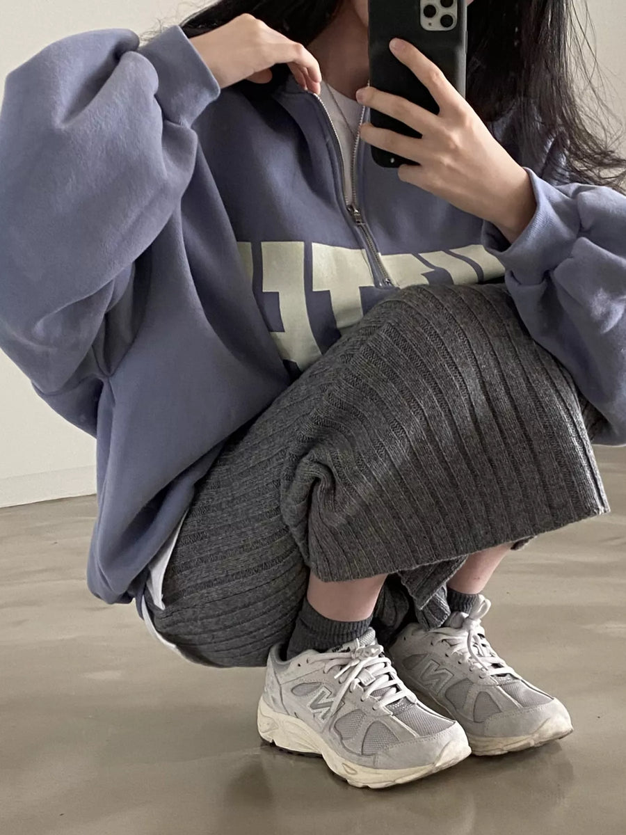 A person crouching in a mirror selfie wearing an oversized dusty purple quarter-zip sweatshirt, a gray ribbed knit skirt, gray socks, and light gray New Balance sneakers.