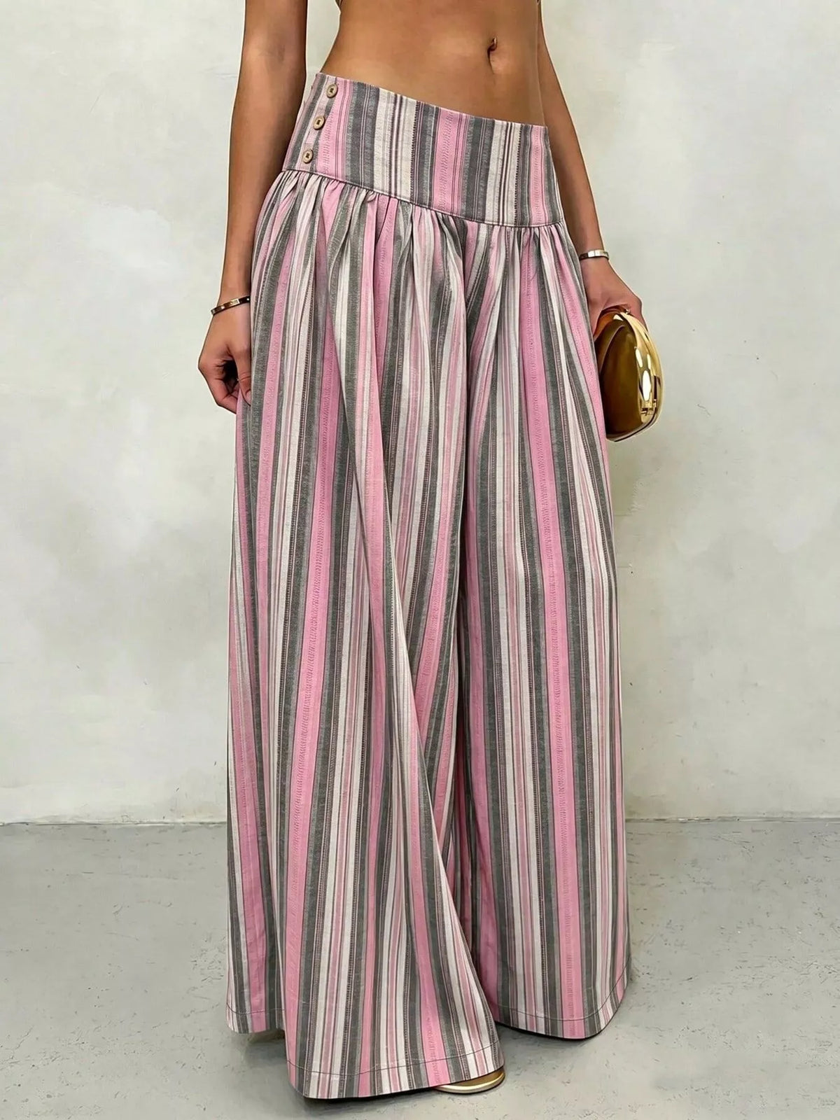 A model wearing high-waisted, wide-leg trousers with vertical pink, gray, and white stripes, holding a small gold clutch against a neutral gray wall.