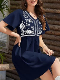 Woman wearing a navy blue dress with white floral embroidery sitting against a wooden wall.