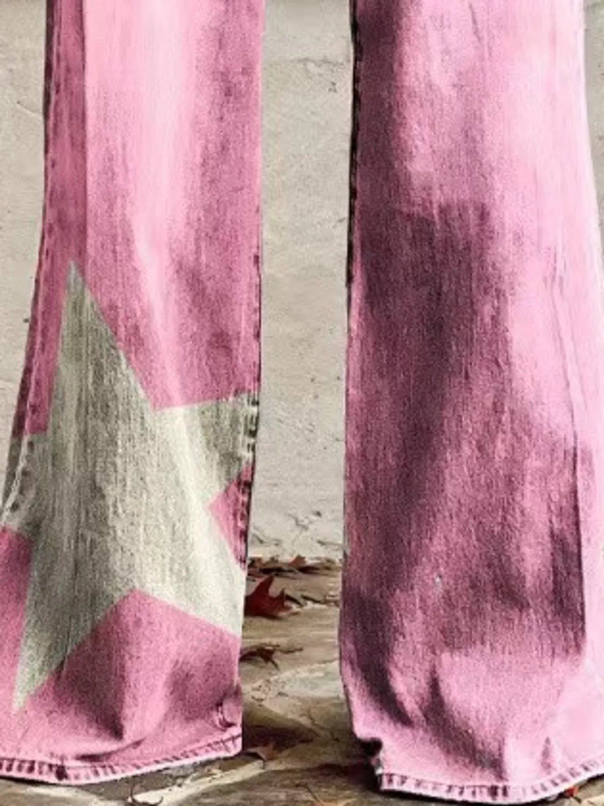 Pink wide-leg denim pants featuring a large distressed gray star graphic on the left leg, shown from the knees down against a rustic stone wall.