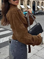 Woman in a brown suede jacket holding a black handbag on a city street.