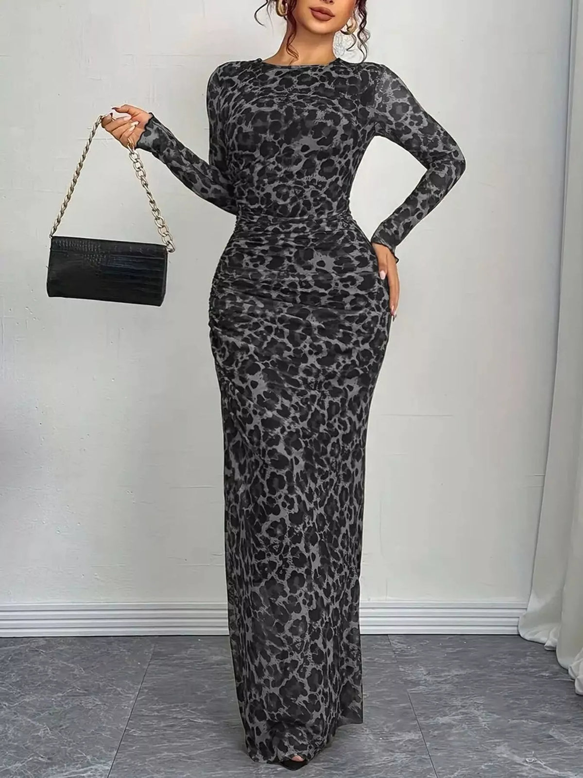 Woman wearing a gray leopard print dress holding a black handbag against a white wall.