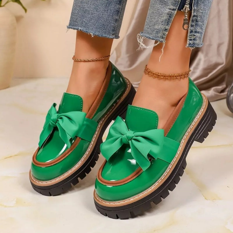 A pair of women's green patent leather loafers with oversized satin bows and chunky lug soles, shown on a model's feet with frayed blue jeans.