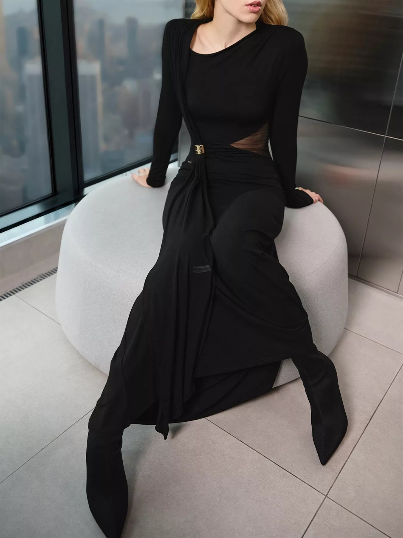 Person wearing a black outfit sitting on a white stool with a cityscape view in the background.