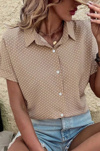 Woman wearing a beige polka dot shirt and denim shorts against a stone wall.