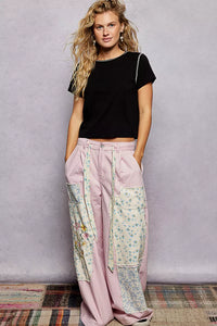 Woman wearing a black top and floral patchwork pants against a gray wall.