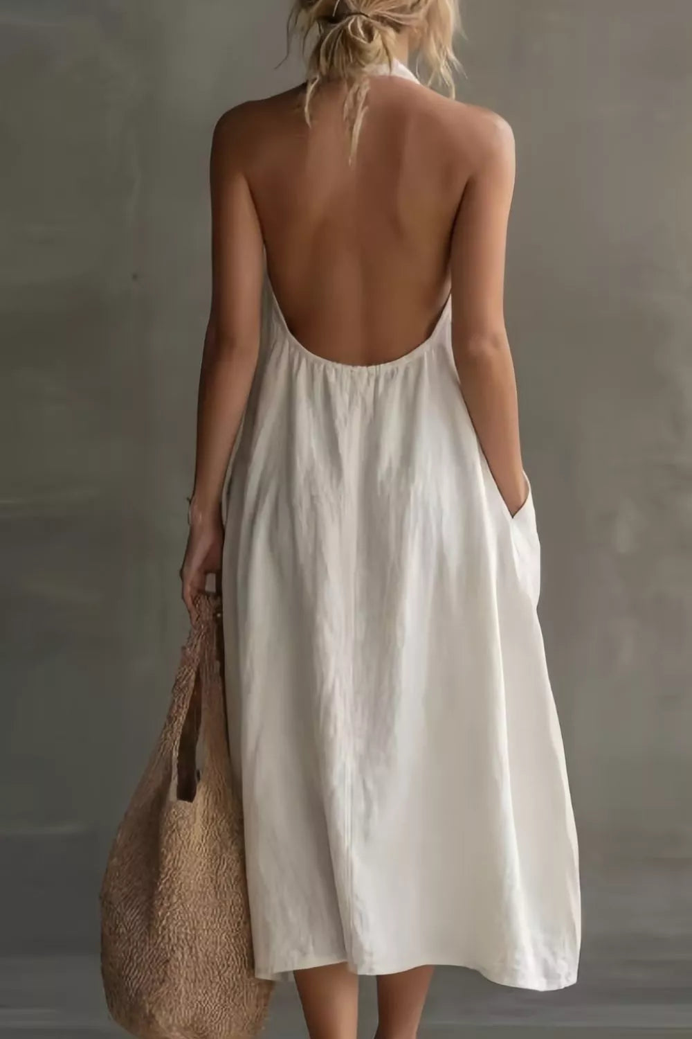 Woman wearing a white dress with an open back, holding a woven bag against a neutral background
