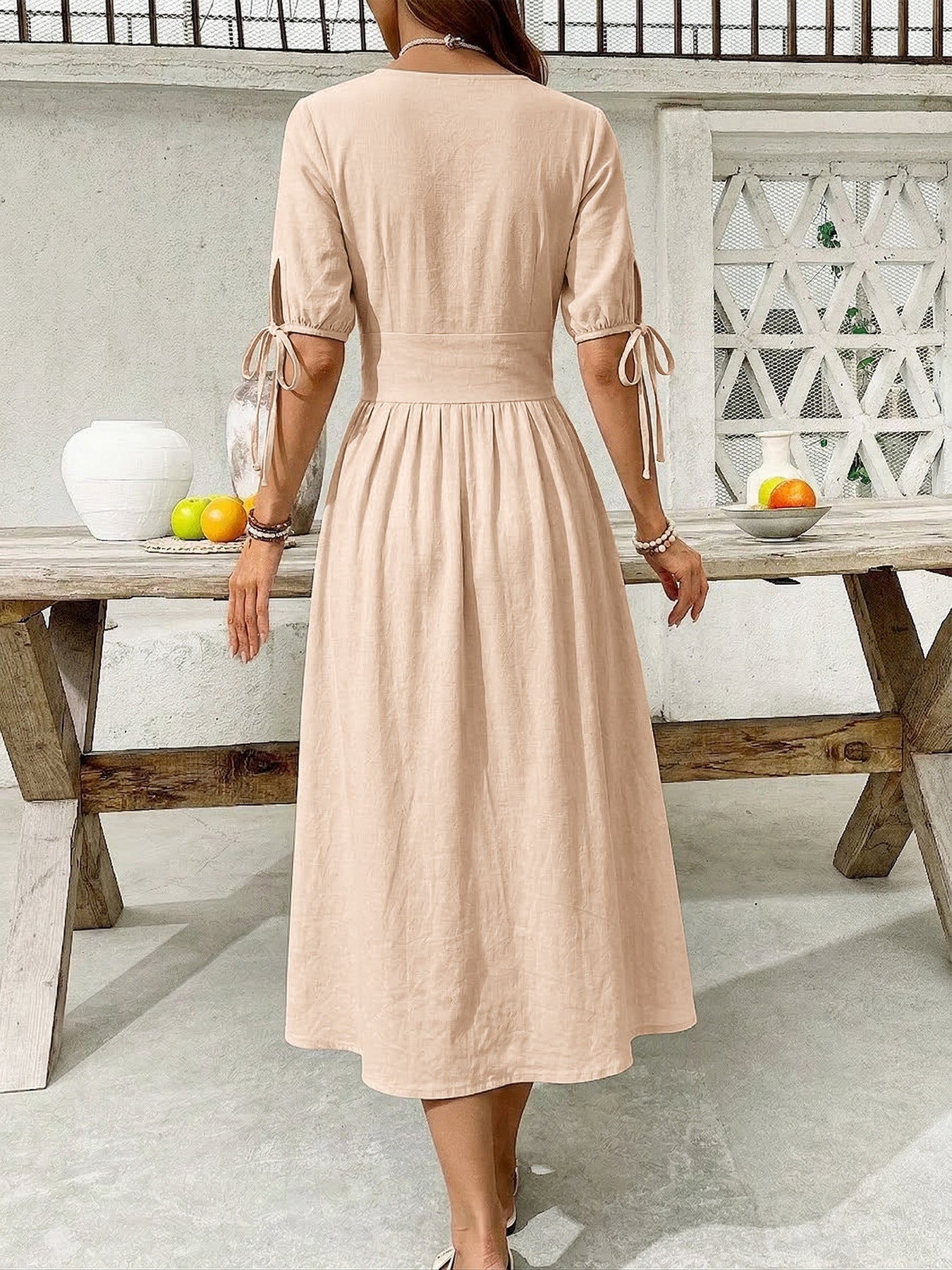 Woman in a beige dress standing by a wooden table outdoors.