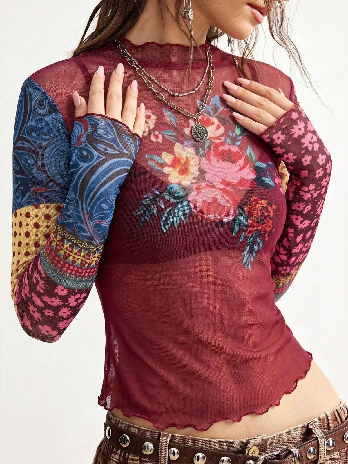 A woman wearing a sheer long-sleeve top with a burgundy base, colorful floral print on the chest, and patterned sleeves in blue, pink, and yellow, paired with brown studded pants.