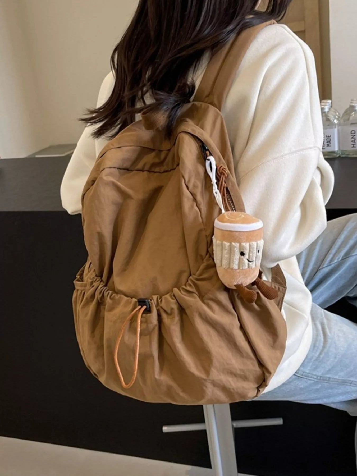Person wearing a brown backpack with a plush toy attached, sitting indoors.