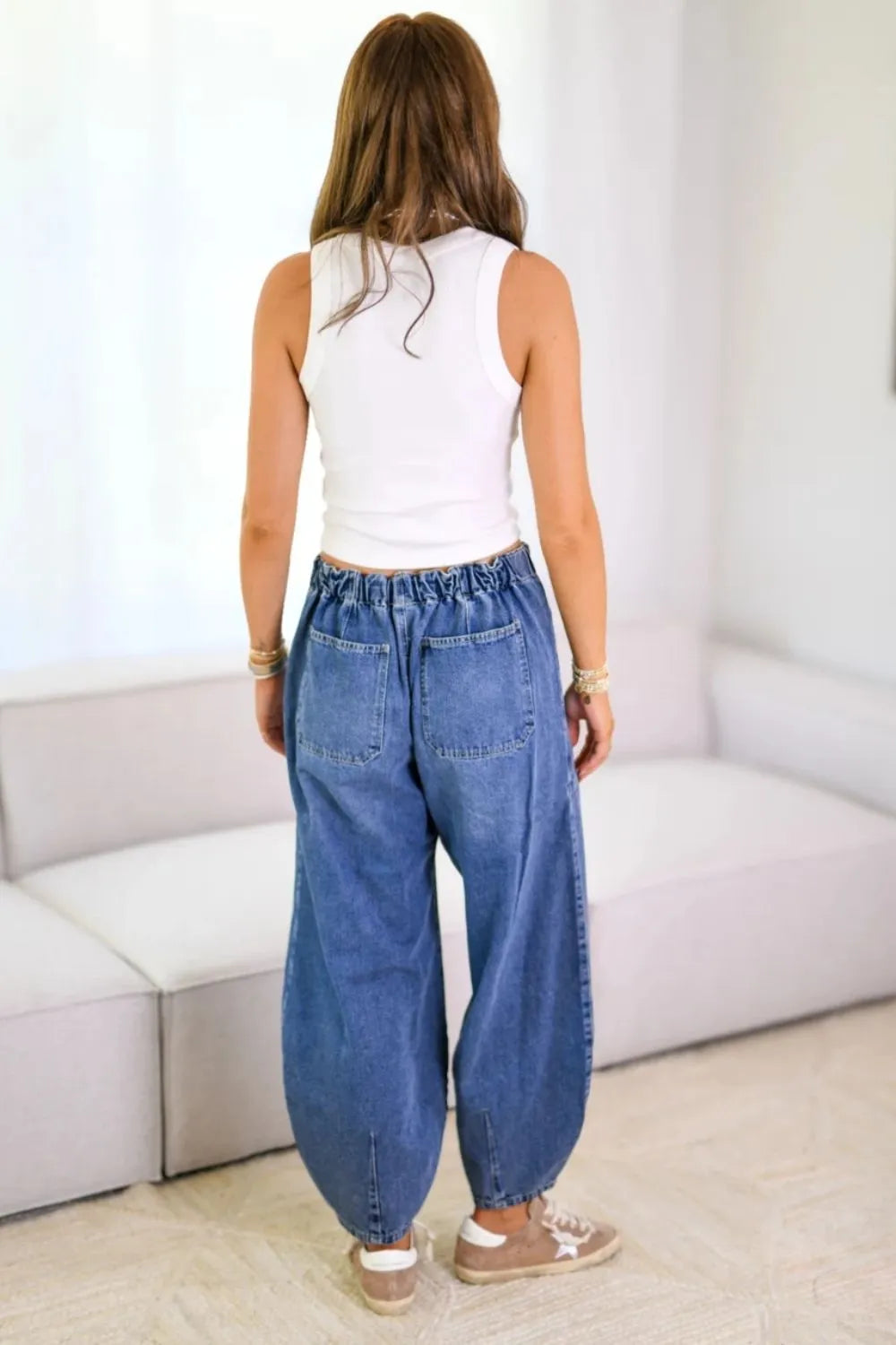 Person wearing a white sleeveless top and blue jeans standing in a minimalistic room.