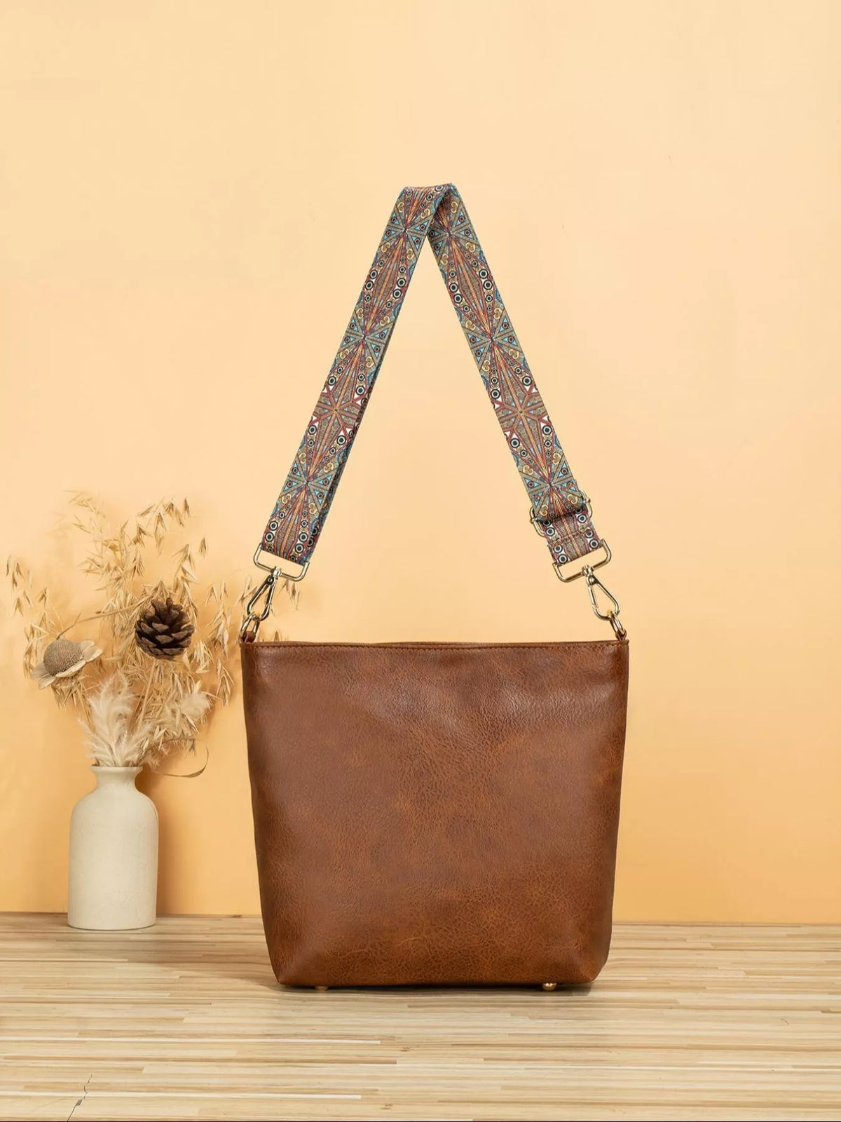 Brown leather handbag with a patterned strap on a wooden surface and beige background