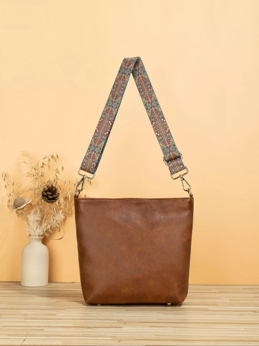 Brown leather handbag with a patterned strap on a wooden surface and beige background