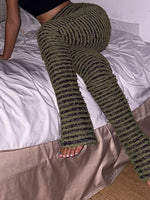 A person wearing ribbed, textured knit pants in olive green and black, sitting on a bed with white sheets and a beige slipcover.