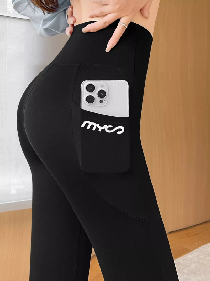 Black high-waisted athletic leggings with a side pocket holding a silver smartphone, featuring the white stylized 'mxc2' logo on the pocket. The model is shown from the waist down in an indoor setting with a light wood door in the background.