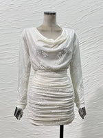 An off-white long-sleeve mini dress with a draped cowl neckline and subtle floral embroidery, displayed on a mannequin against a textured white wall.