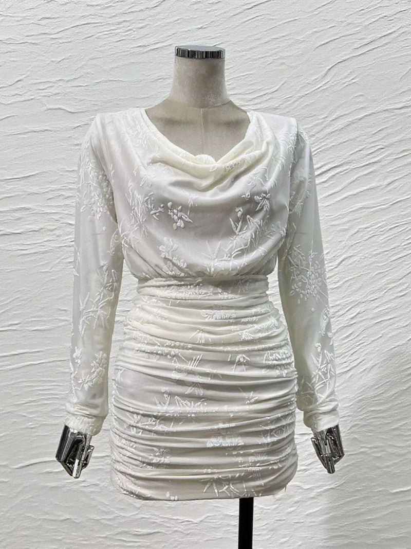 An off-white long-sleeve mini dress with a draped cowl neckline and subtle floral embroidery, displayed on a mannequin against a textured white wall.