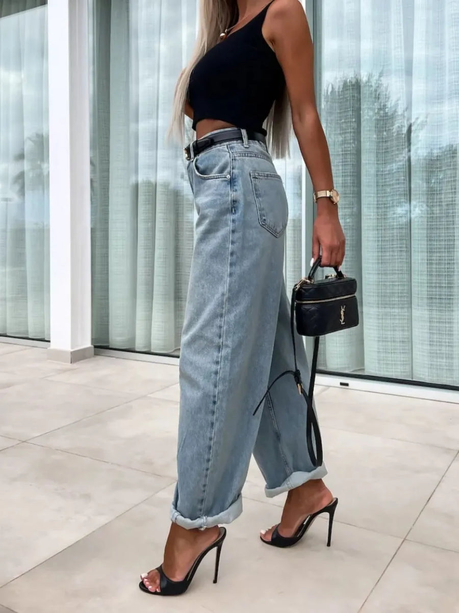 Woman wearing a black top and light blue jeans, holding a black handbag indoors.
