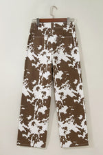 Brown and white abstract patterned pants hanging on a wooden hanger against a plain light beige wall.