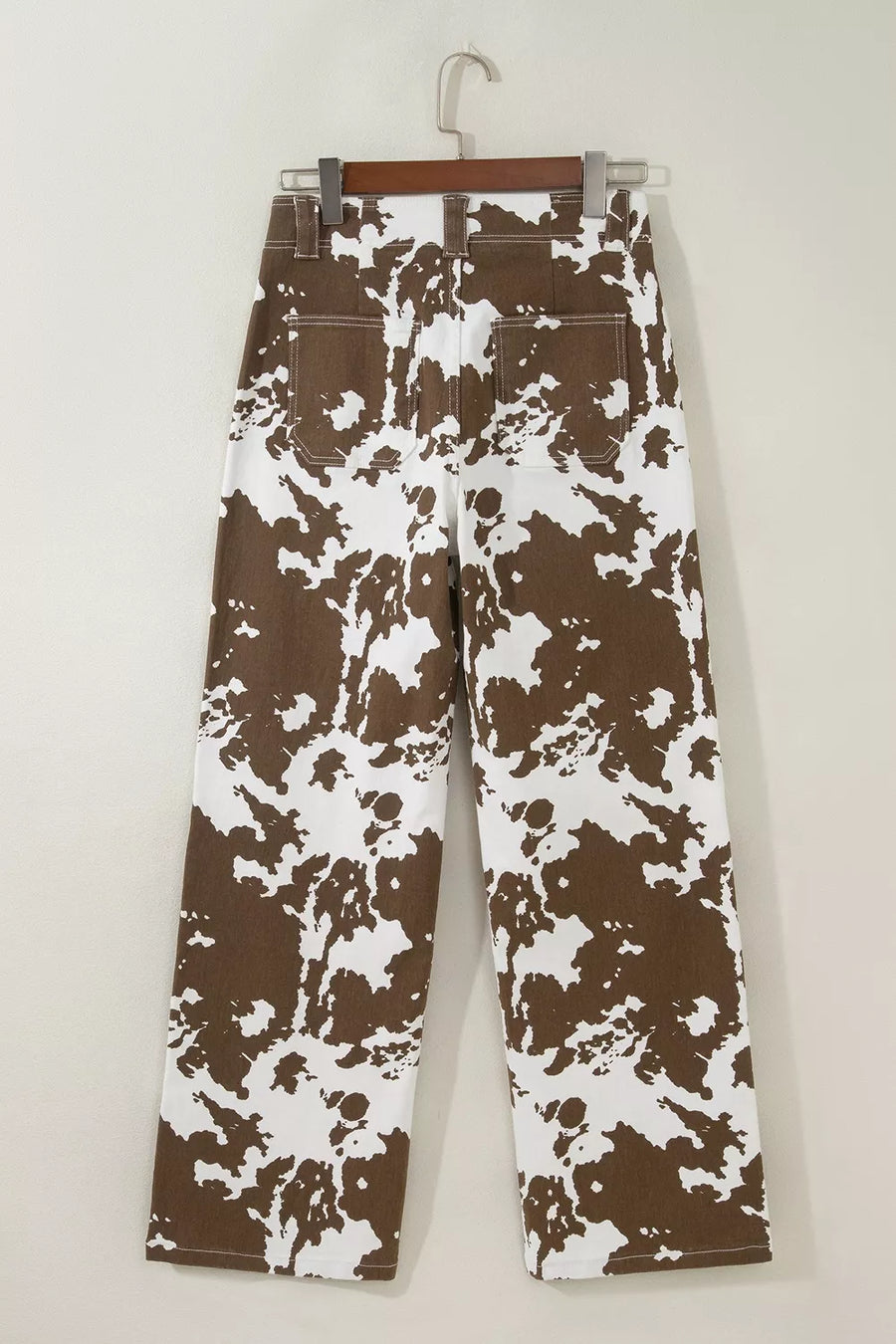 Brown and white abstract patterned pants hanging on a wooden hanger against a plain light beige wall.