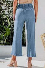 Person wearing light blue jeans standing outdoors with plants and a white wall in the background