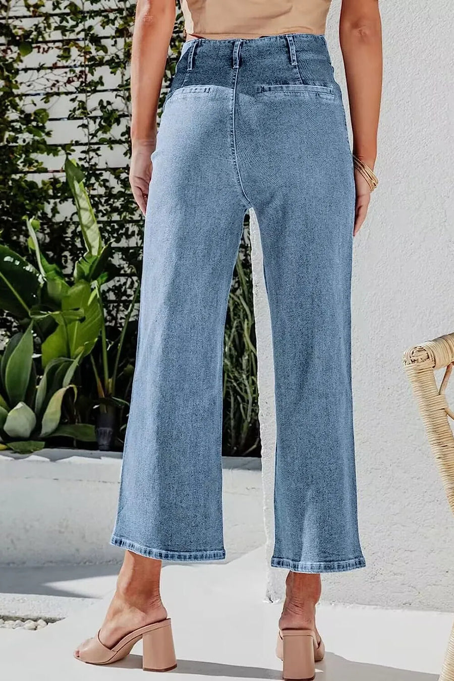 Person wearing light blue jeans standing outdoors with plants and a white wall in the background