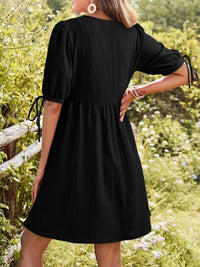 Person wearing a black dress standing in a natural setting with greenery.