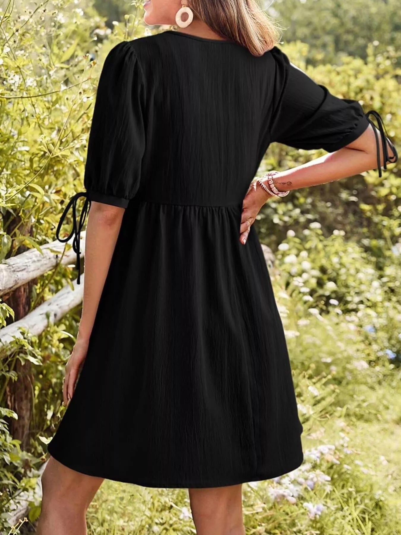 Person wearing a black dress standing in a natural setting with greenery.