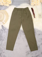 A pair of olive green slim-fit trousers laid flat on a white faux fur surface with decorative props including a straw hat, flowers, a brown belt, and a small purse.