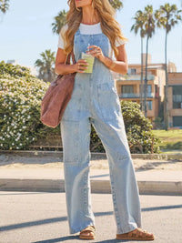 Woman wearing light blue denim overalls standing outdoors with palm trees and buildings in the background.