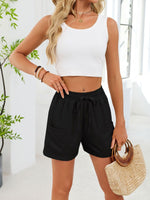 Woman wearing a white tank top and black shorts, holding a straw bag indoors.