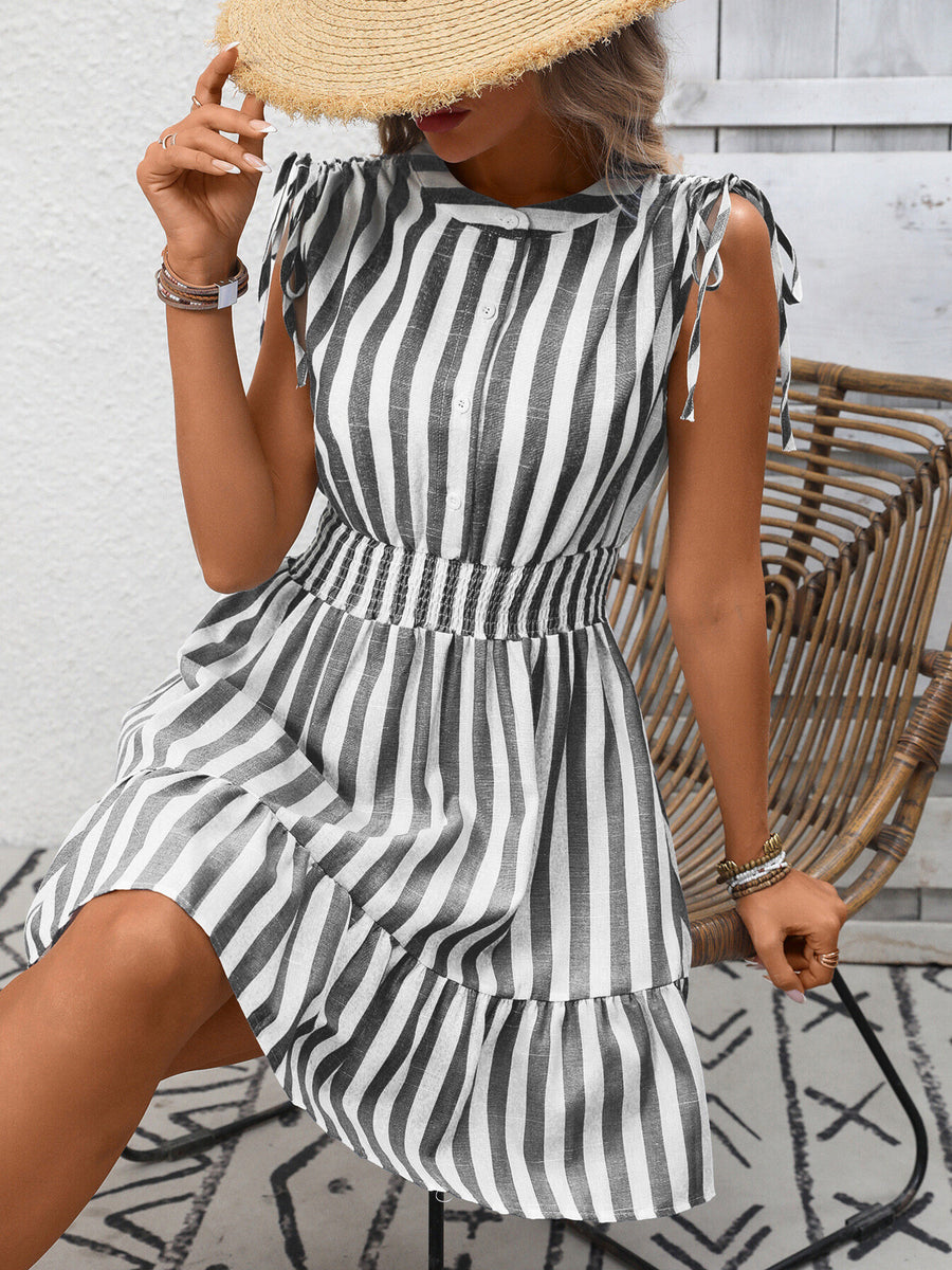 Woman wearing a black and white striped dress with a straw hat, standing against a light wooden wall.