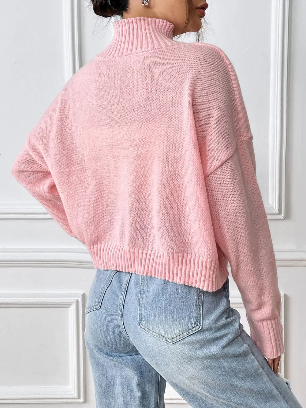 Person wearing a pink sweater and light blue jeans against a white wall.