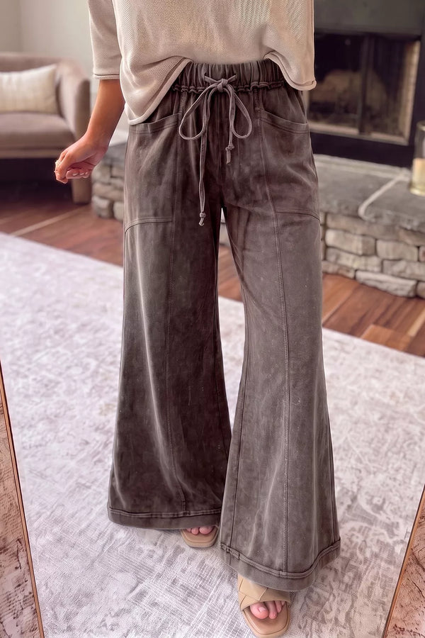 A person wearing wide-leg charcoal gray drawstring pants and a light beige top, standing indoors on a patterned rug in front of a fireplace.