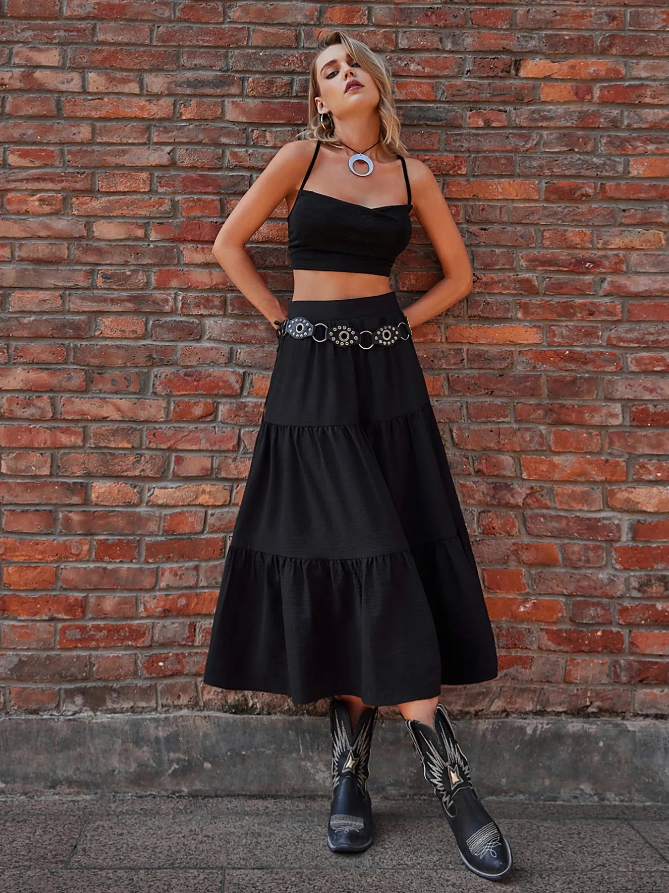 Woman in a black outfit with a belt standing against a brick wall.