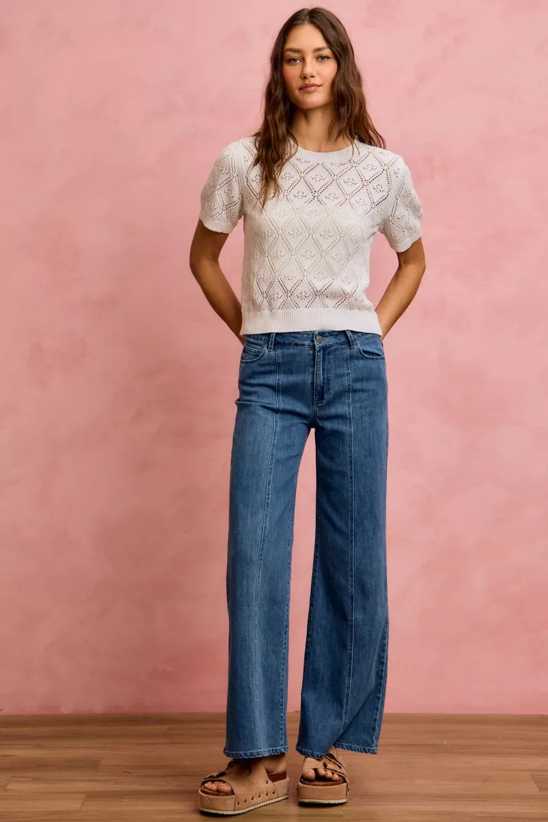 Woman wearing a white lace top and blue jeans against a pink wall.
